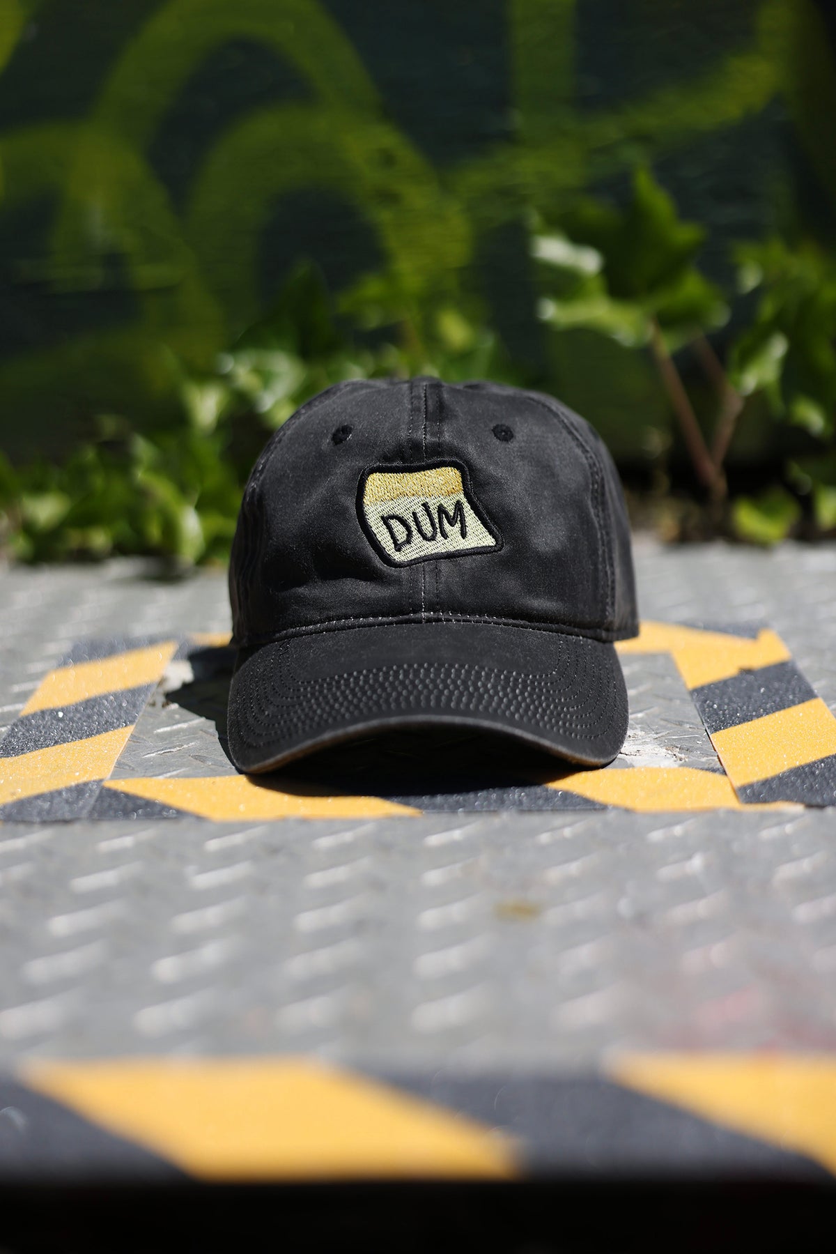 A close up photograph of the black DUM hat on a metal grate in front of a graffitied wall background.