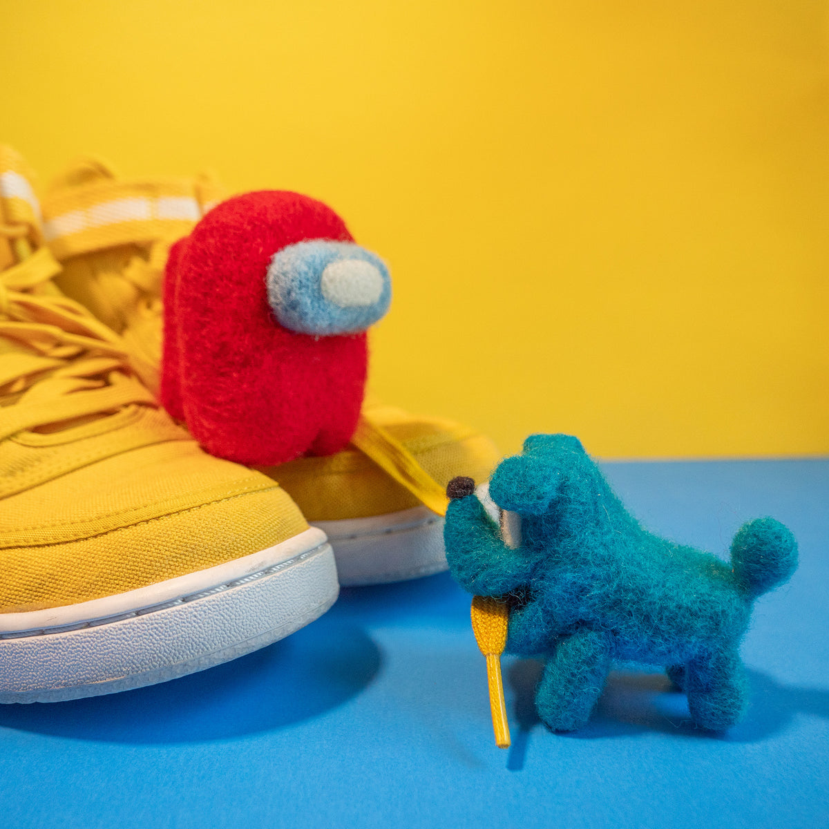 The felted crewmate and spacedog standing on a pair of yellow sneakers. The crewmate is watching the spacedog, who has the shoelace in its mouth.