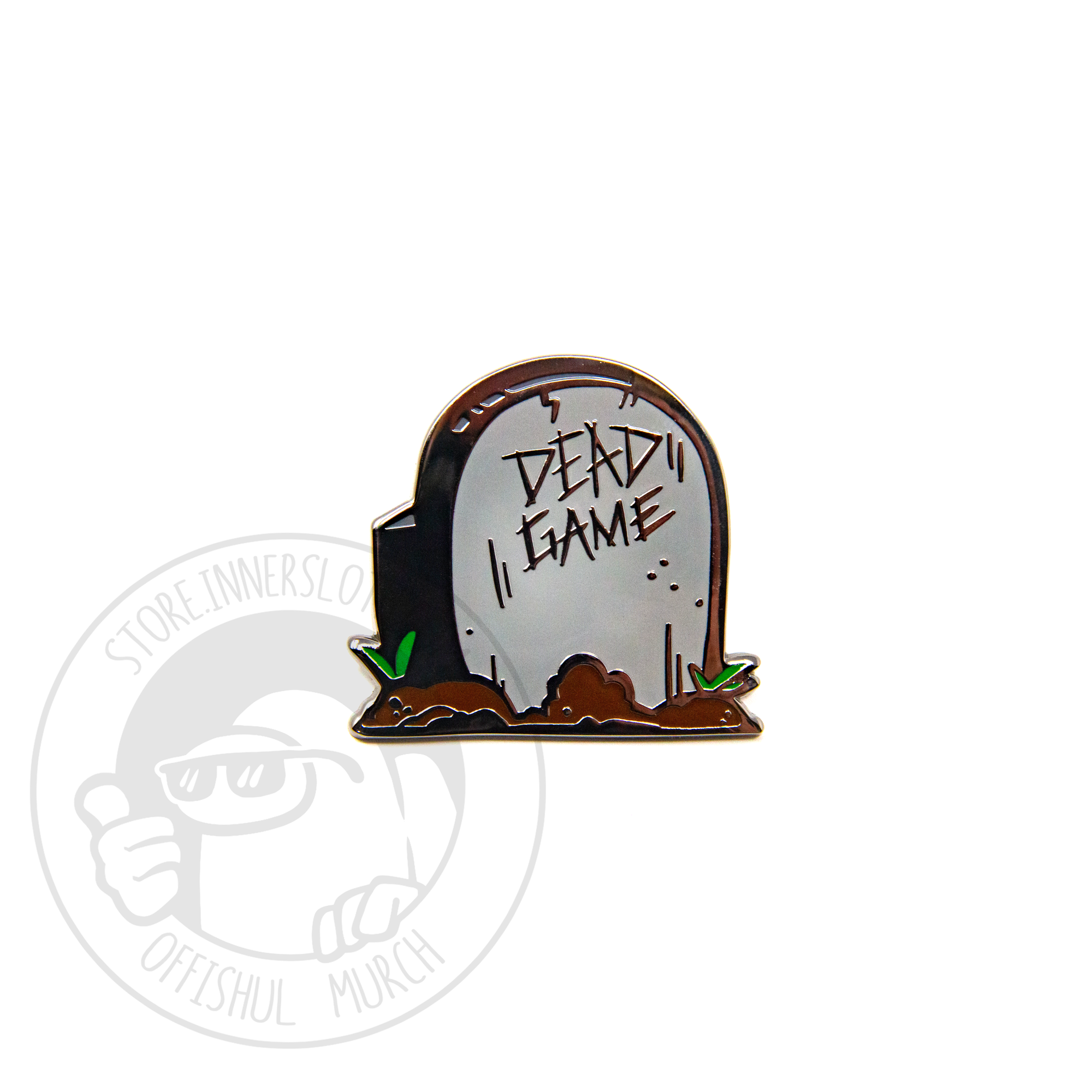 A product photo of a hard enamel pin depicting a grey headstone surrounded by small mounds of fresh dirt and grass sprouts. The headstone is vaguely crewmate-shaped, and reads “DEAD GAME.”