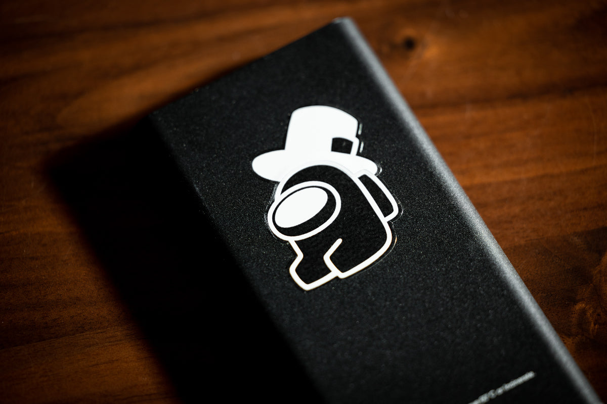 A close up photograph of a white outlined sitting Crewmate with a top hat stuck to the back of a cell phone.