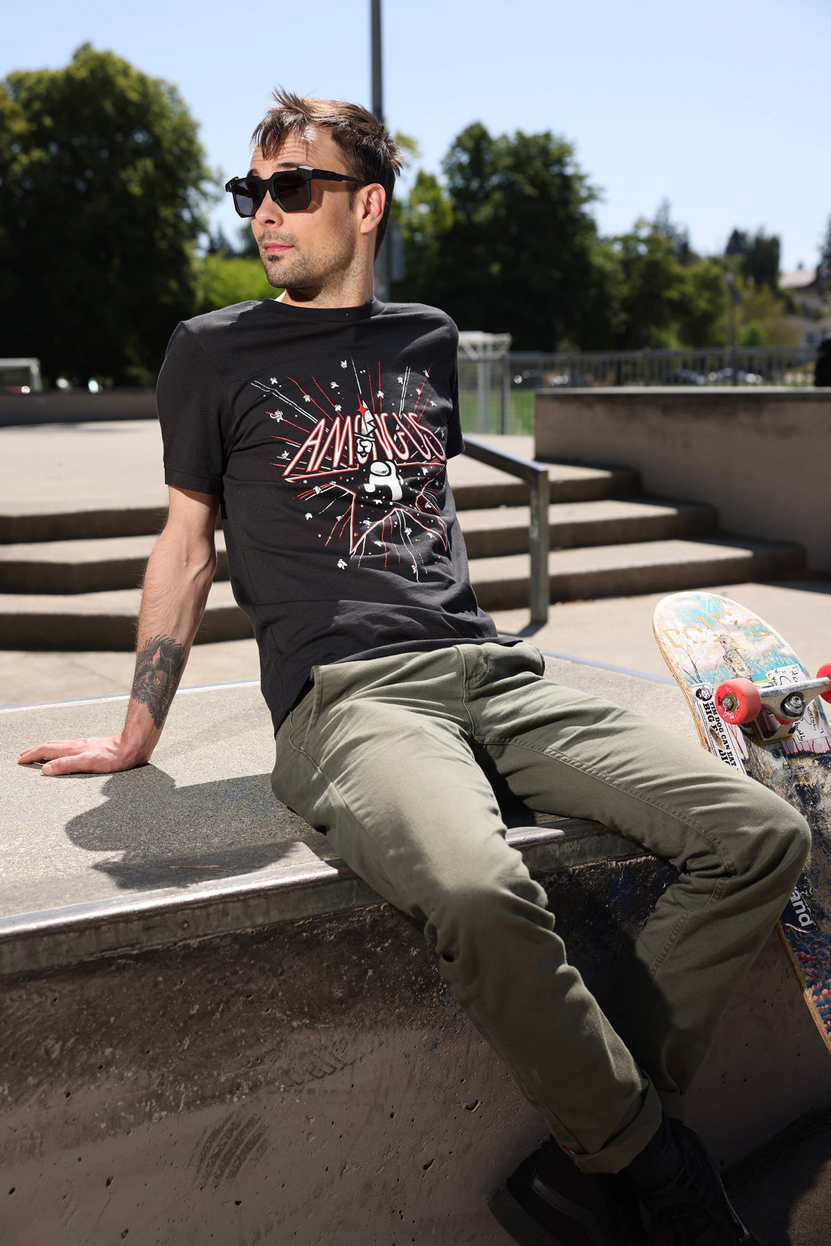 A nearly full-body photograph of a model wearing the Among Us: 5th Anniversary Tee. The model is sitting on a skateboard ramp looking over his shoulder. 