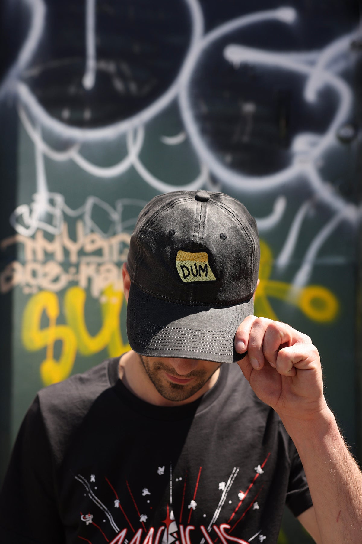 A photograph of the top of a model’s head showing off the brown DUM hat. Their chin is tilted downward so you can see the full view of the yellow “DUM” sticky note embroidery.