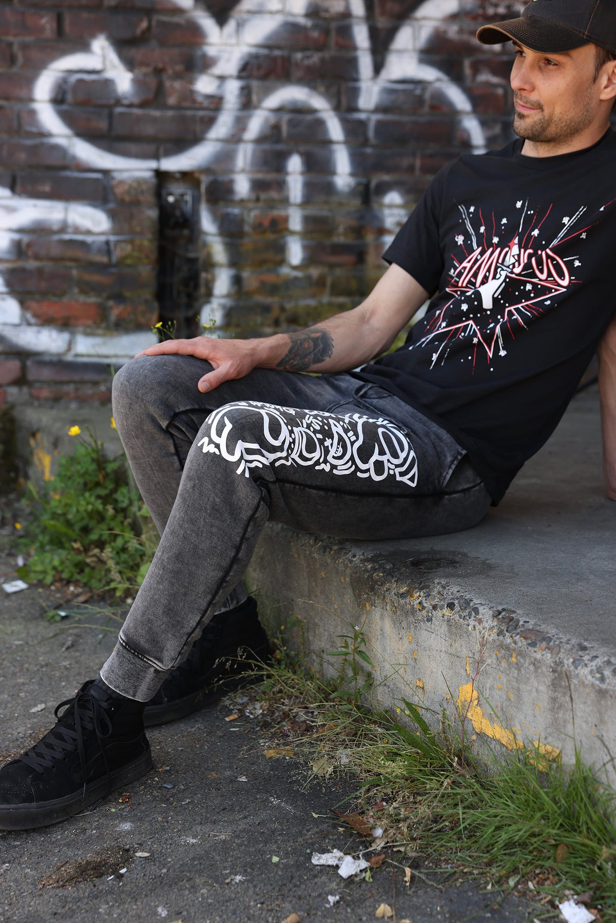 A 3/4, full body, photograph of the model sitting on a step. They are wearing the DUM hat by Puffballs United, the 5th Anniversary Tee by Amy Liu, and the Crewmate Joggers by Kiri Krogsgaard. 