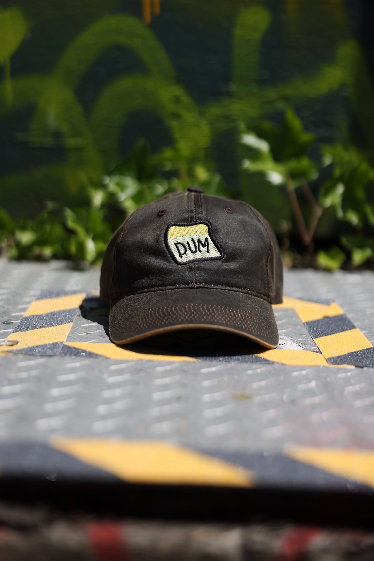 A close up photograph of the brown DUM hat on a metal grate in front of a graffitied wall background.