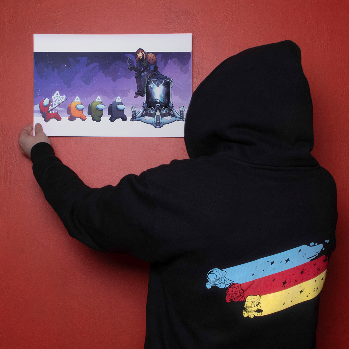  A Model holding up the Bank the Motes Task print against a red wall. The model’s back is turned toward the viewer showing off the  Eyes Up, Crewmate Hoodie screenprint. 