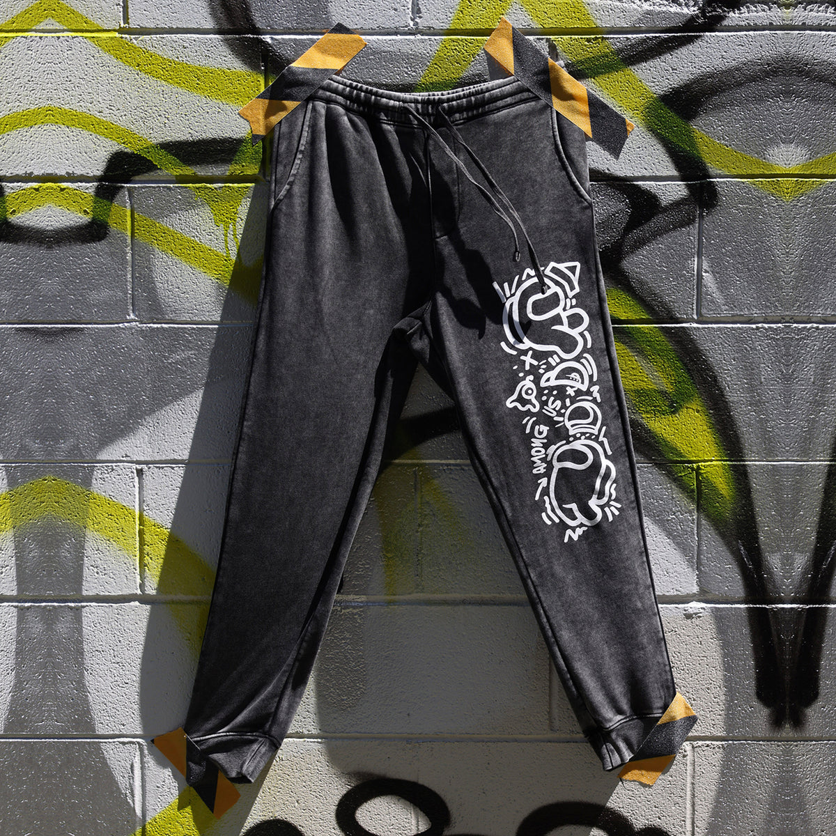 A photograph of the gray joggers laid flat and taped to a graffitied wall. The joggers are being held to the wall by yellow and black caution tape.