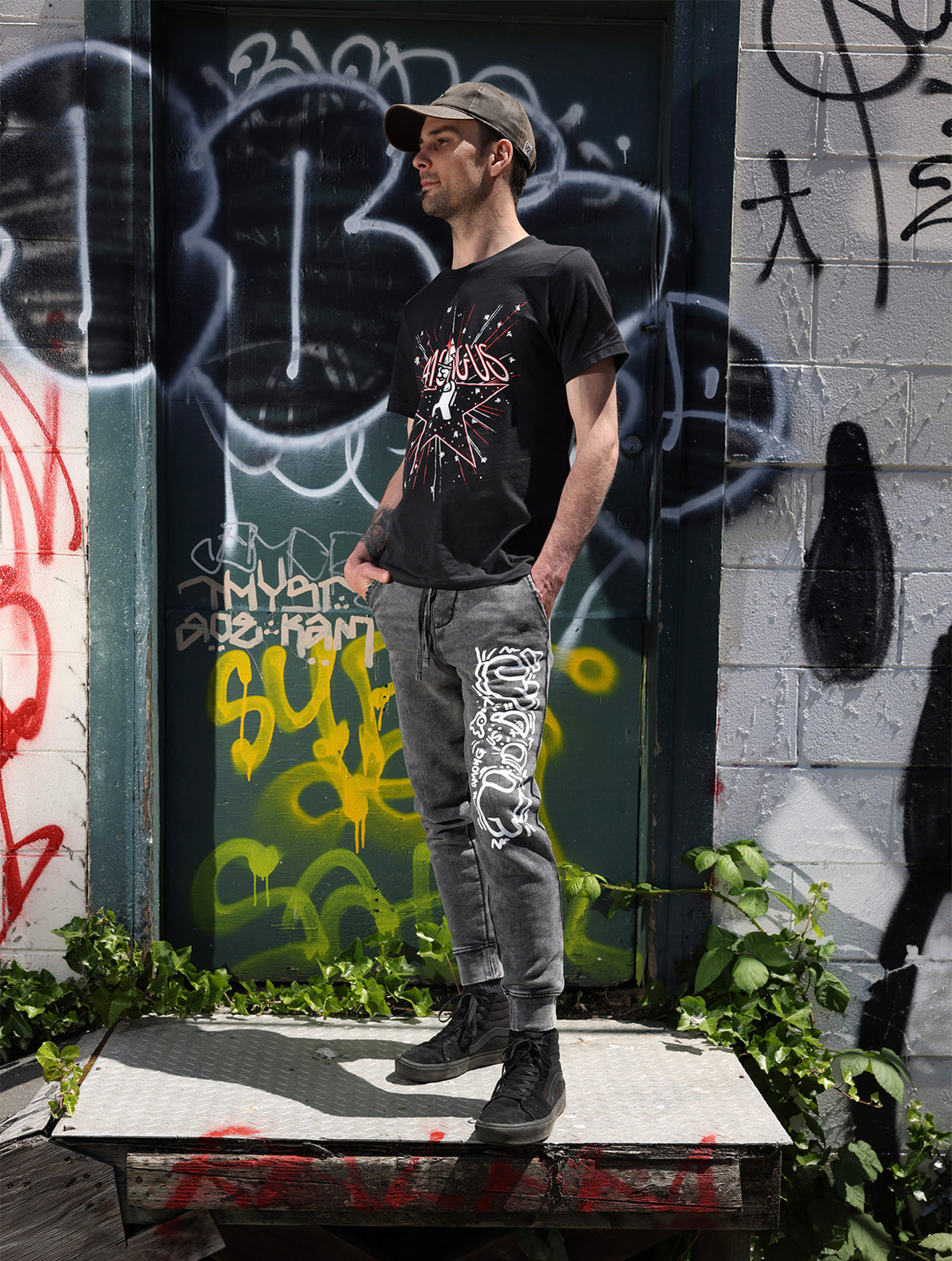  A ¾ full body photograph of a model standing on a grate against a graffitied background. He’s wearing the DUM hat designed by Puffballs United, the Among Us: 5th Anniversary Tee designed by Amy Liu, and the Crewmate Joggers designed by Kiri Krogsgaard. 