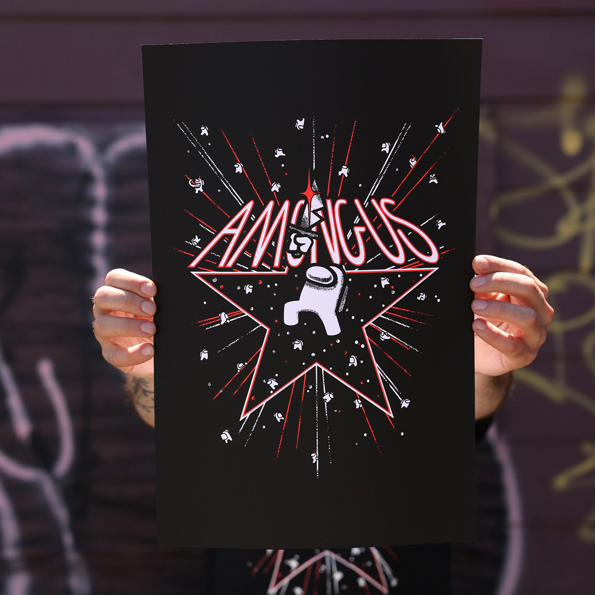 A close up photograph of a model’s hands holding up the Among Us: 5th Anniversary Print  by Amy Liu. The print depicts a white Impostor among a flurry of stars and fireworks. The impostor stands in the middle of a red outlined star holding a bloody knife over their head. “Among Us” text is written in red and white sitting on top of the outlined star. 