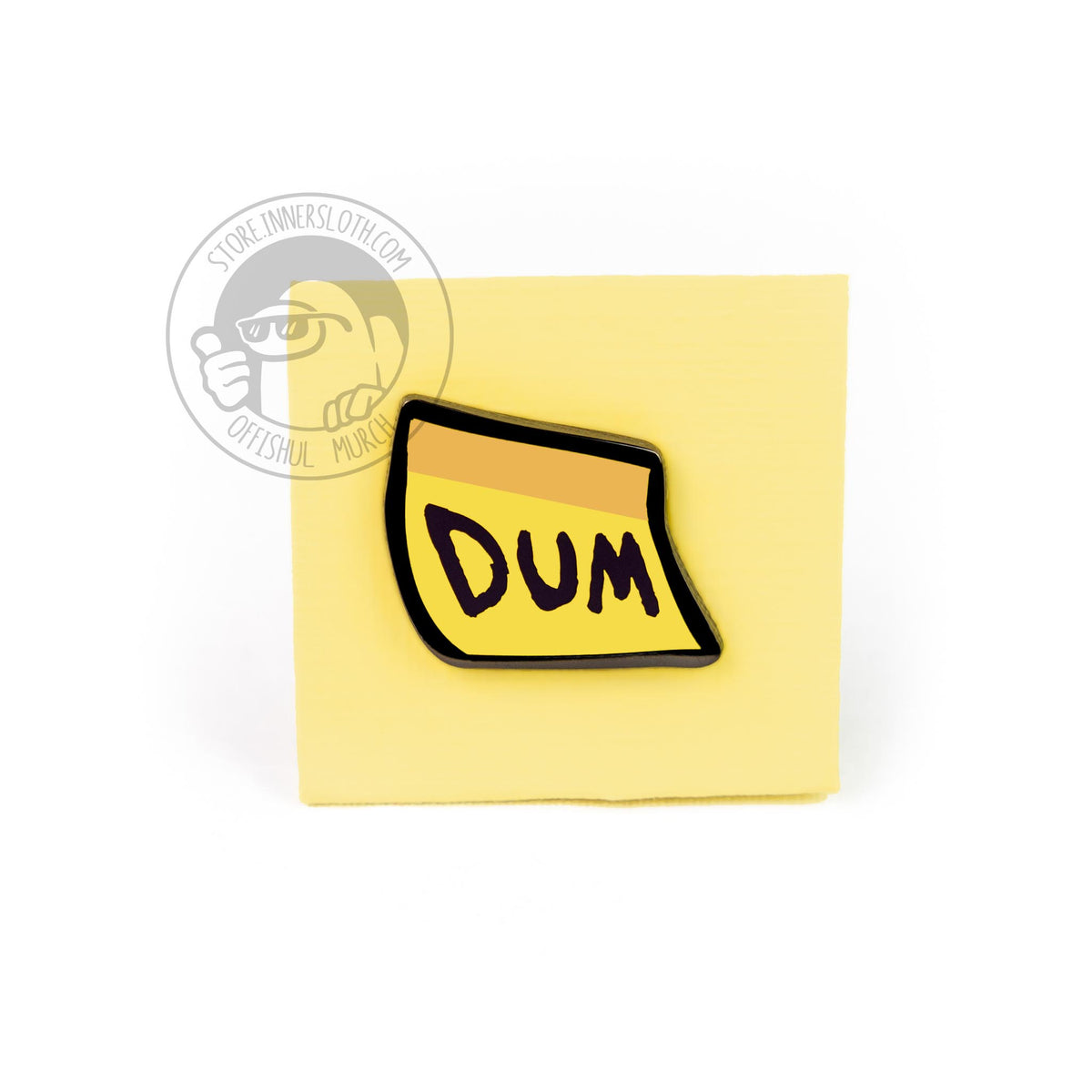 The DUM sticky-note enamel pin, pinned to a real paper sticky note on a white background.