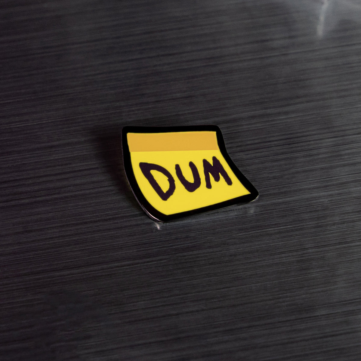 An enamel pin of the yellow sticky-note visor cosmetic on a metal table. DUM is written in large letters across the front of the note.