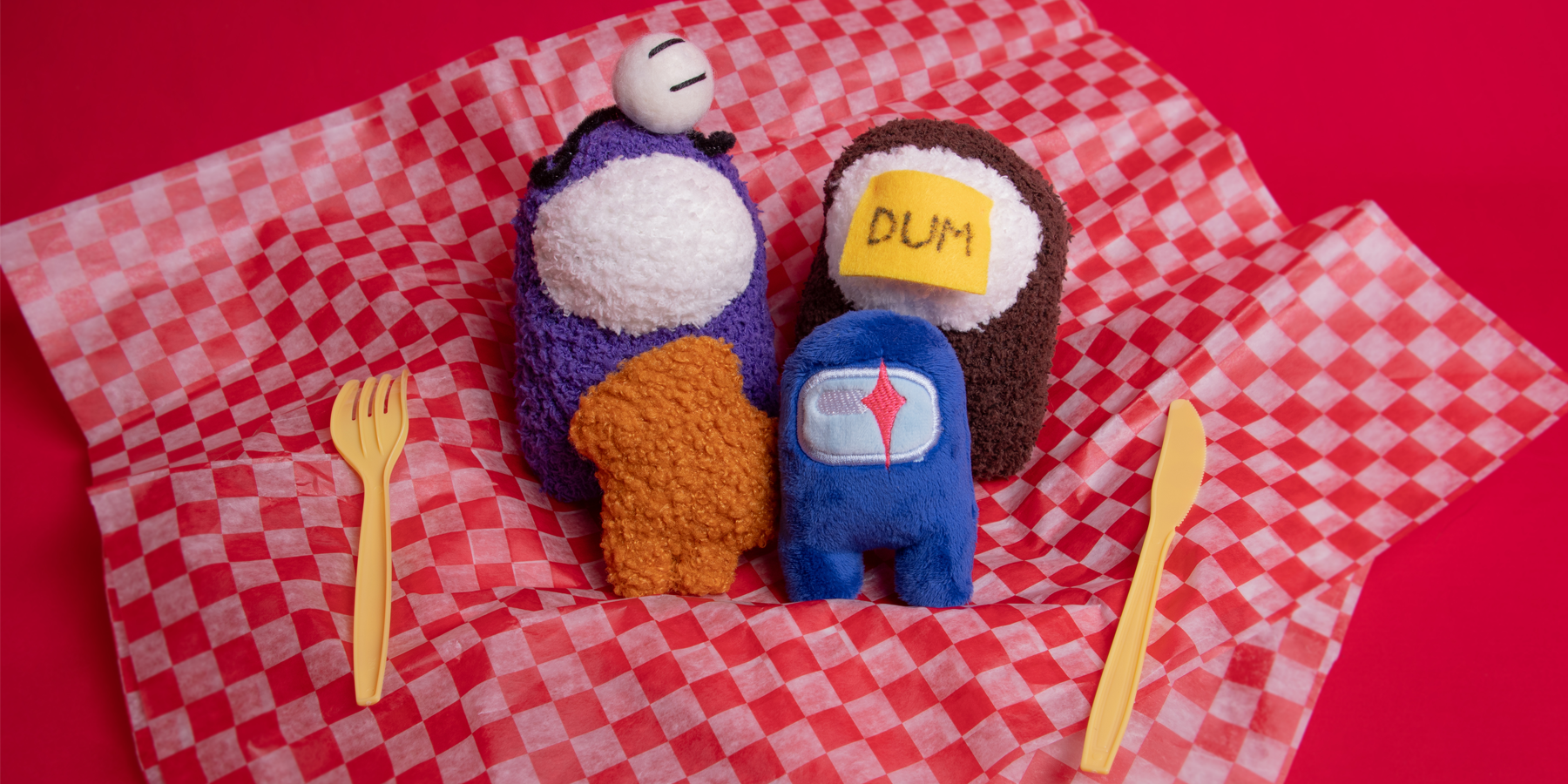 A grouping of four Crewmate plushies in the middle of a sheet of checkered diner tissue paper. There is a yellow plastic fork and knife on either side of the plush.