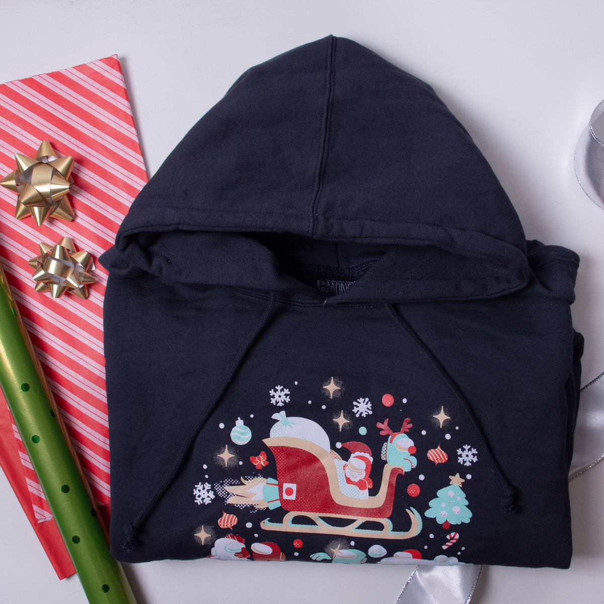 A photograph of the black Christmas Crewmates hoodie folded atop a striped piece of wrapping paper laid next to bows, tissue paper, and a roll of wrapping paper. 