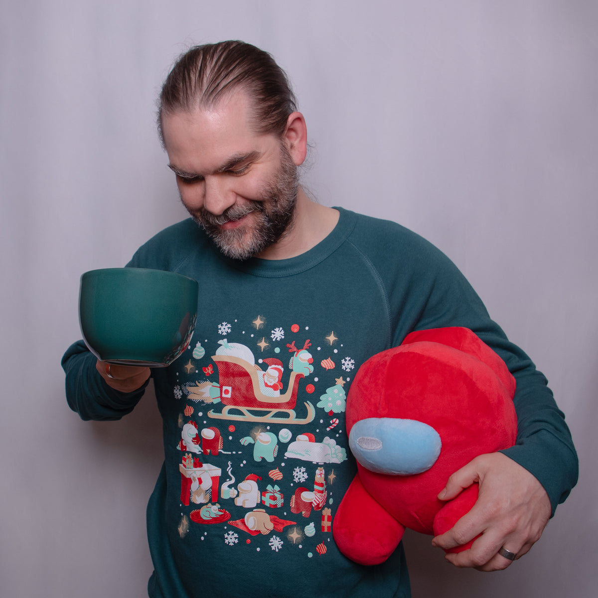 A photograph of a model wearing a green Christmas Crewmates longsleeve sweatshirt. The model is holding up a dark green mug in one hand and holding a red Crewmate plush in the other.