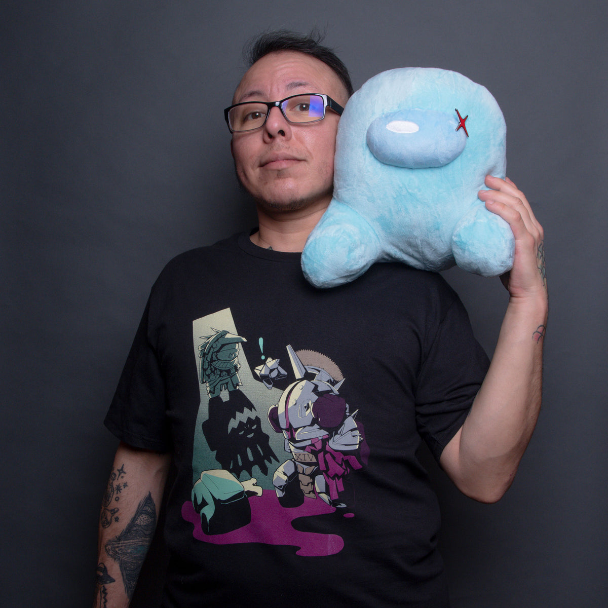 A model wearing the Osiris the Impostor tee holding a Cyan Sitting Crewmate Plush on their shoulder. The plush has an Impostor Shine pin pinned to the left side of its visor. 
