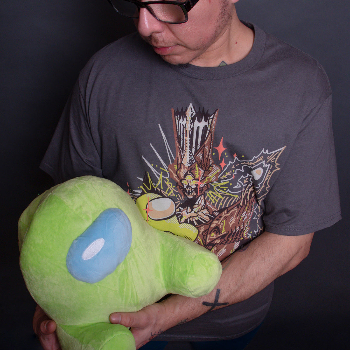  An overhead photo of a model wearing the Witch Bean Tee. The model looks down at a Lime Green Crewmate Plush held against the bosom of the shirt.
