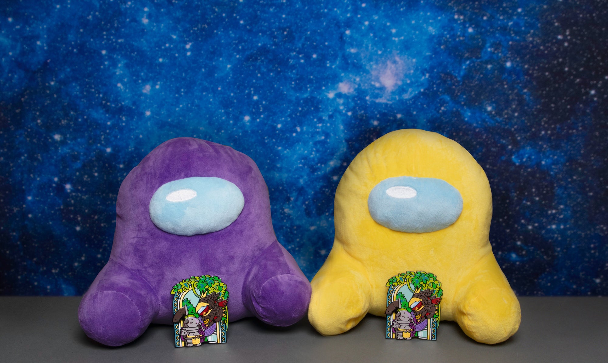 A Purple and Yellow Crewmate plush sitting against a starry, blue, gradient background. Each Plush has a Among Us x Destiny 2: Peace to Purpose Enamel Pin propped up in front of them.