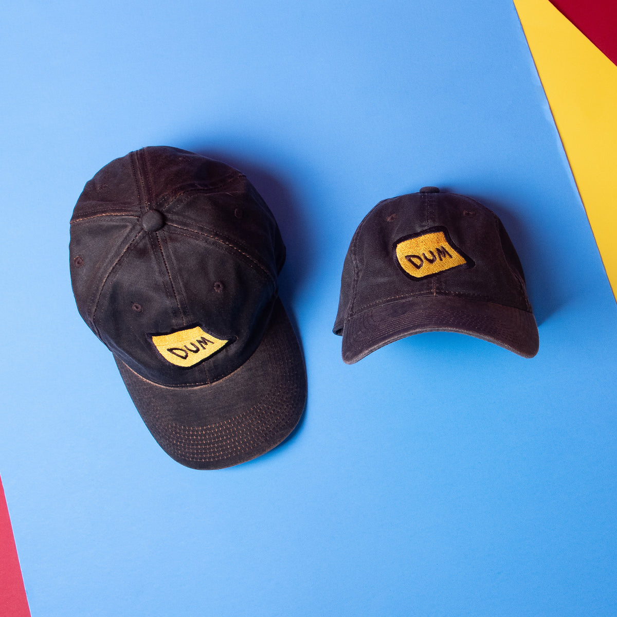 An overhead photograph of two Black DUM hats. One hat shows the entire cap from above and the hat next to is tilted upward so you can see the full view of the embroidery and brim on a red, yellow, and blue color-blocked background.