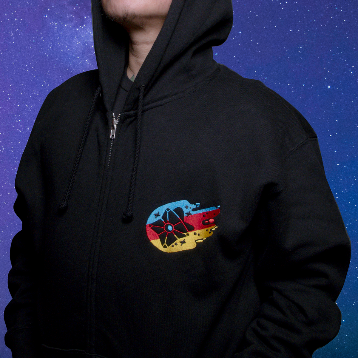 A model standing against a starry blue and purple background wearing a zipped up Eyes Up, Crewmate Hoodie, gazing upward. The colorful blue, yellow, and red embroidered chest hit motif is positioned over the left breast of the garment.