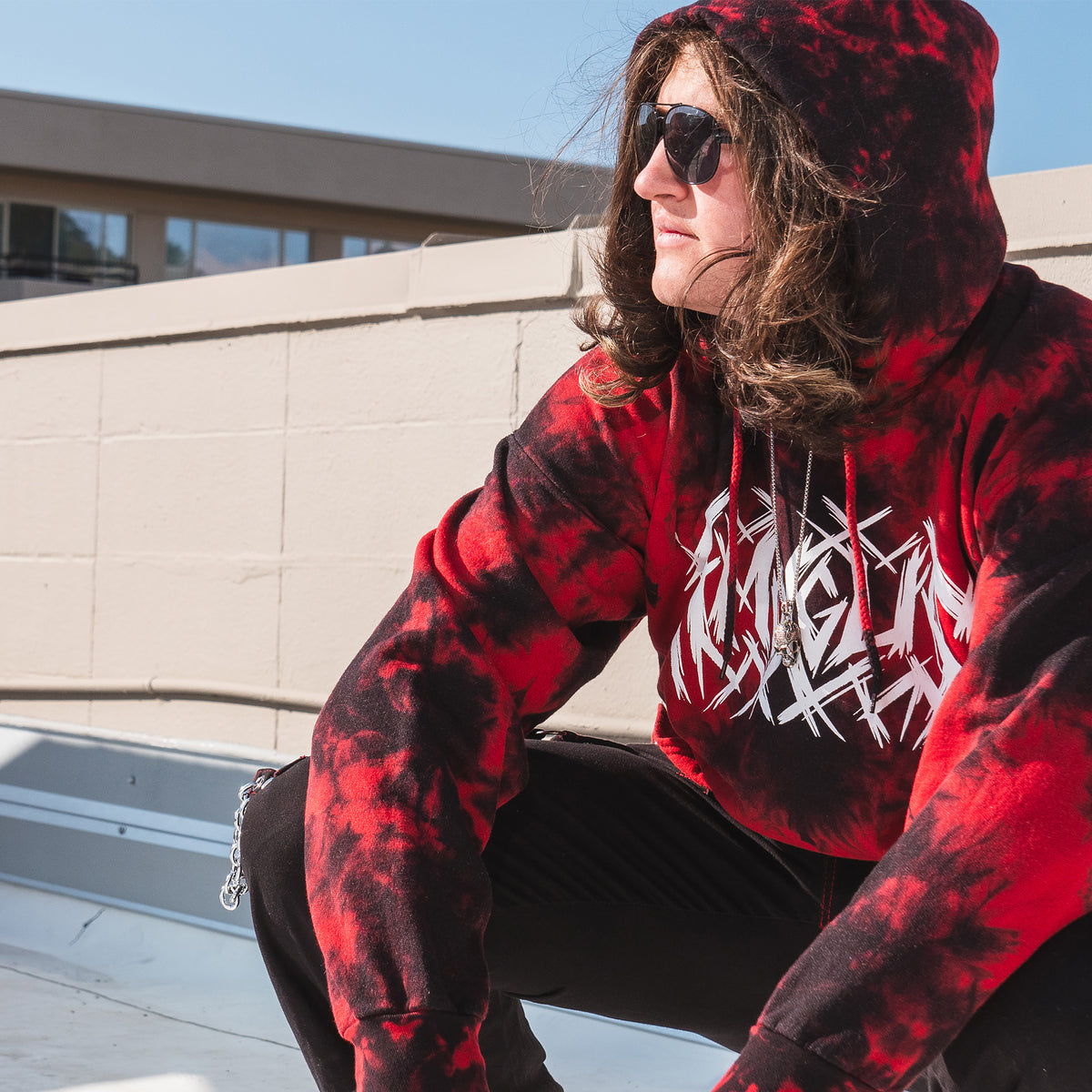 A three-quarter view of a model wearing sunglasses and the hoodie looking left with their arms draped over their knees.The front of the hoodie says “Among Us” in a texture metal music-esque font.