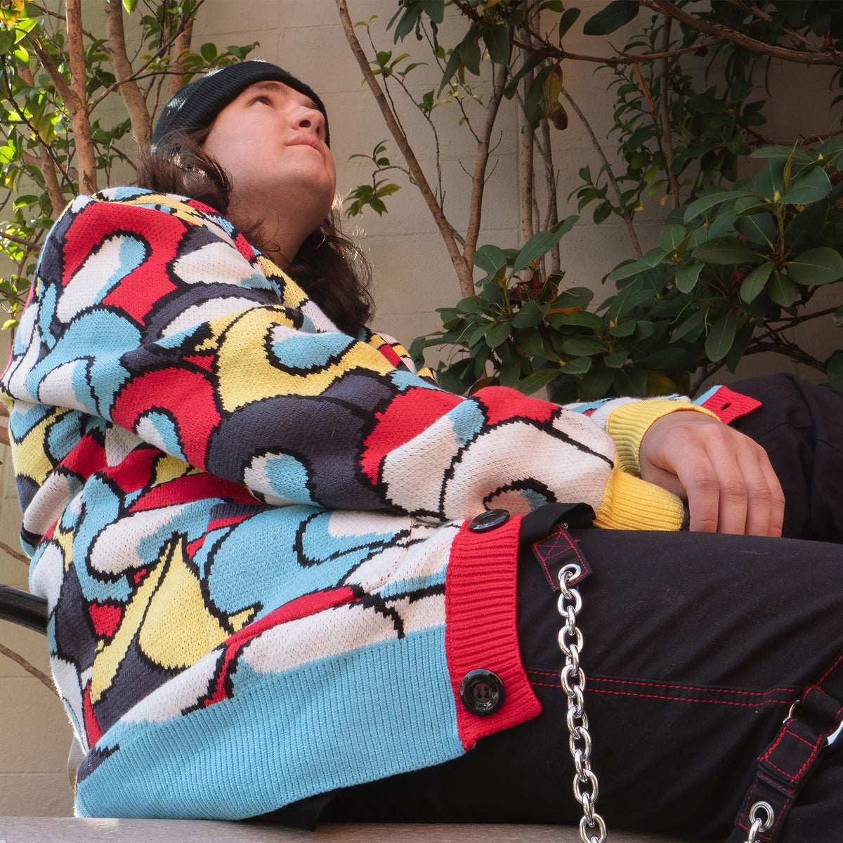 An upward angle shot of a model sitting down, looking up. This shot features a close up of the sleeve and side pattern of the cardigan. 