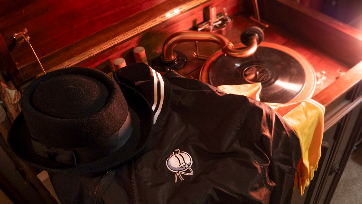  A lifestyle photograph of a black fedora sitting on top of the Henry Stickmin Bomber Jacket. The jacket is draped over a yellow scarf. All the garments are draped over a wooden vintage vinyl record player. The Henry embroidery is shown in full detail over the right breast of the jacket.