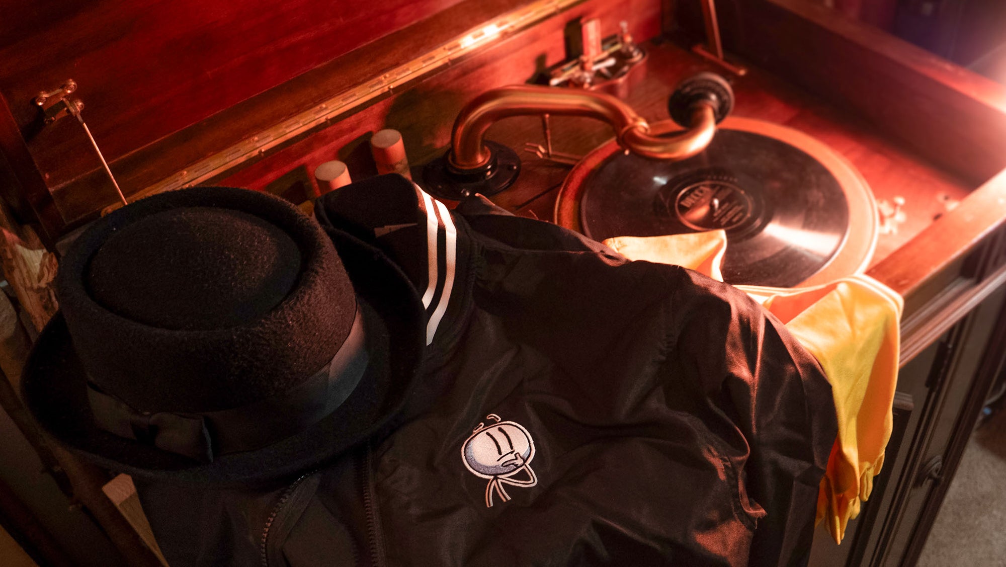 A photograph of a black fedora sitting on top of the Henry Stickmin Bomber Jacket. The jacket is draped over a yellow scarf. All the clothing garments are draped over a vintage vinyl record player. 