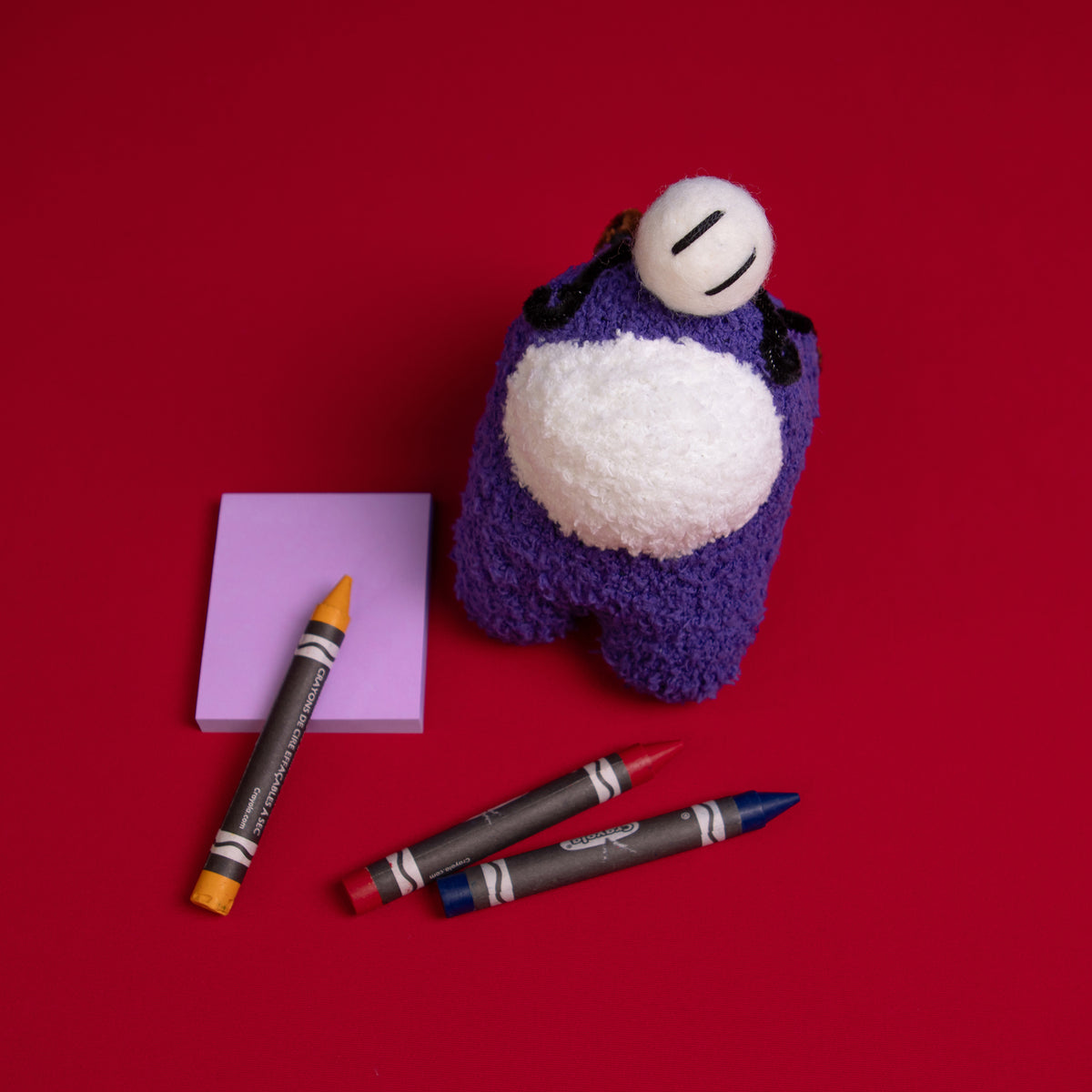 An overhead photograph of the Purple Crewmate and Henry Stickmin plush duo standing to the right of a purple sticky notepad. There is an orange crayon placed over the notepad and a red and blue crayon in front of the Crewmate. 