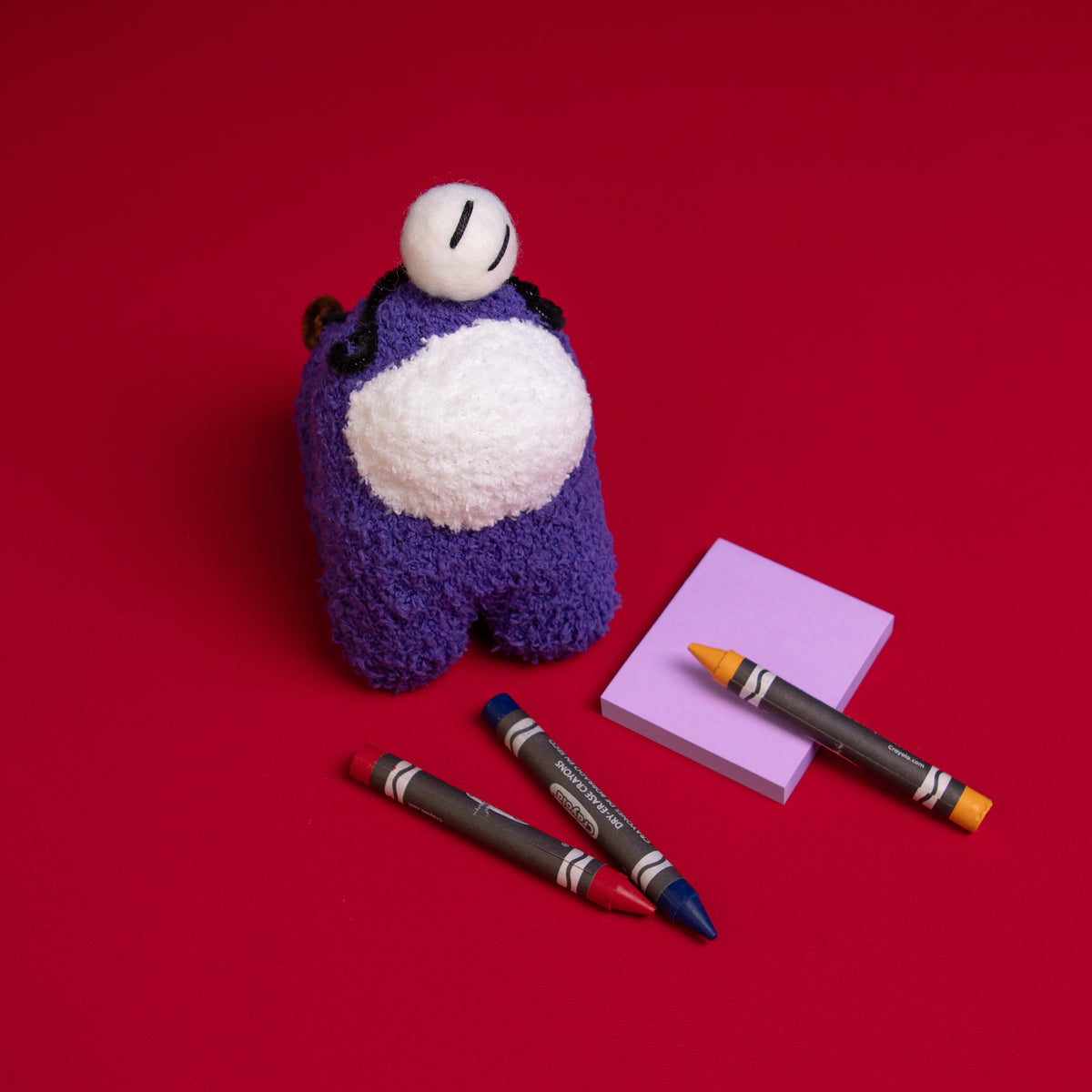 An overhead photograph of the Purple Crewmate and Henry Stickmin plush duo standing to the left of a purple sticky notepad. There is an orange crayon placed over the notepad and a red and blue crayon in front of the Crewmate
