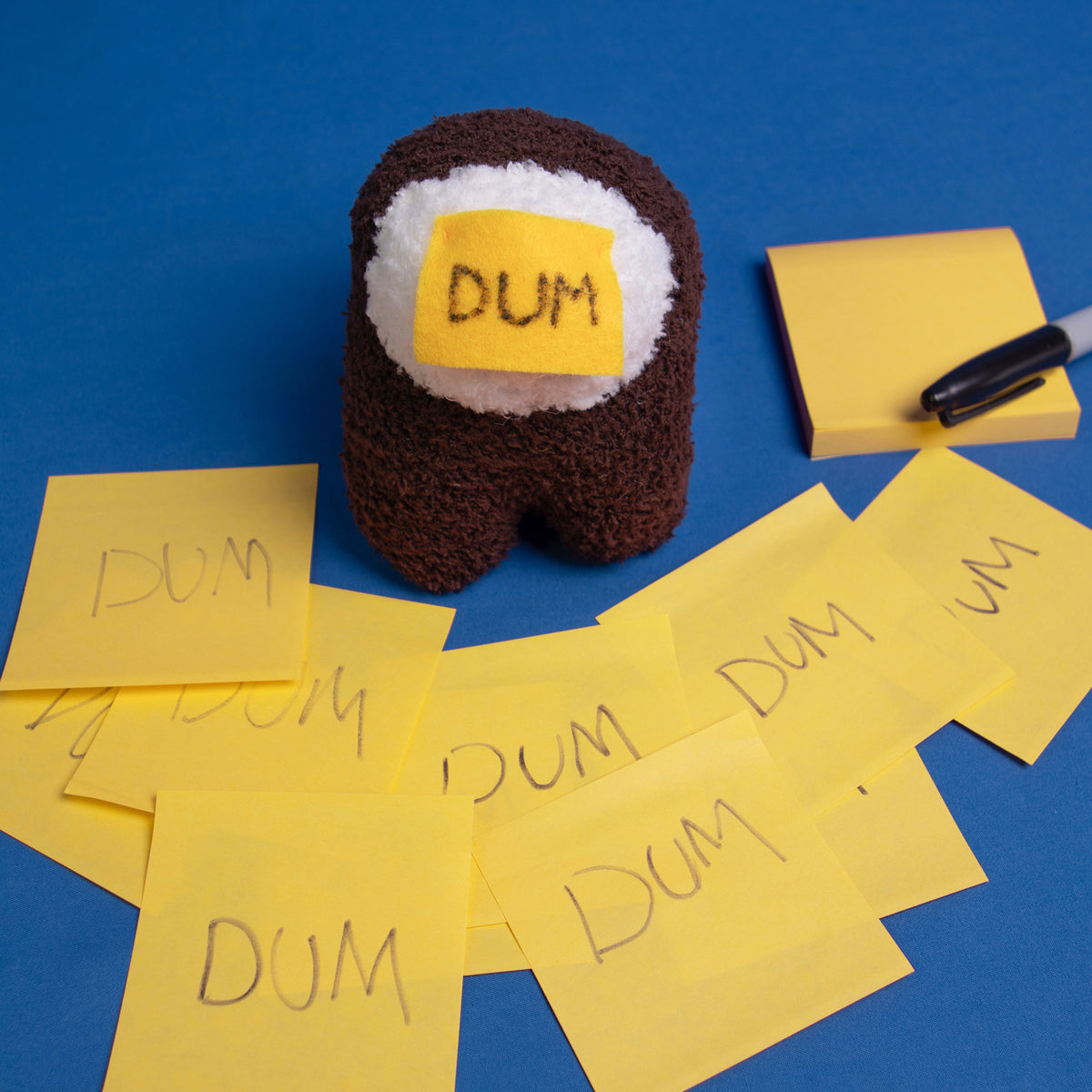 A close up overhead photograph of the DUM brown Crewmate, to the upper right side is a yellow sticky notepad with a permanent marker resting on it. In front of the Crewmate, there are several layered yellow sticky notes that all have DUM written on them