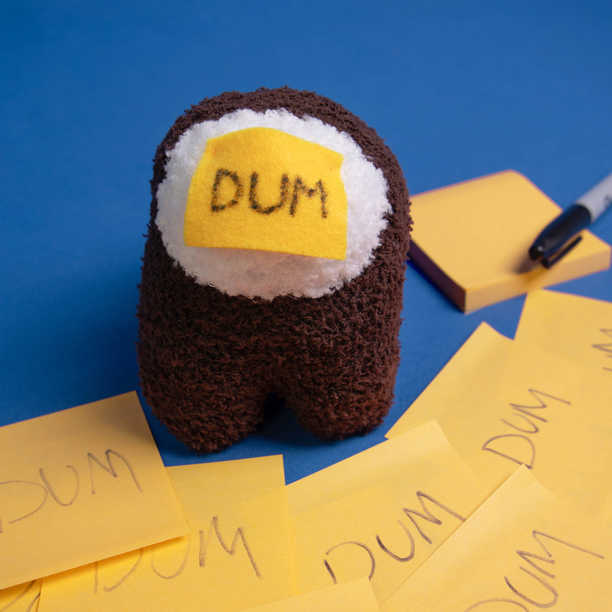 A zoomed in close up of the DUM brown Crewmate, to the upper right side is a yellow sticky notepad with a permanent marker resting on it. In front of the Crewmate, there are several layered yellow sticky notes that all have DUM written on them