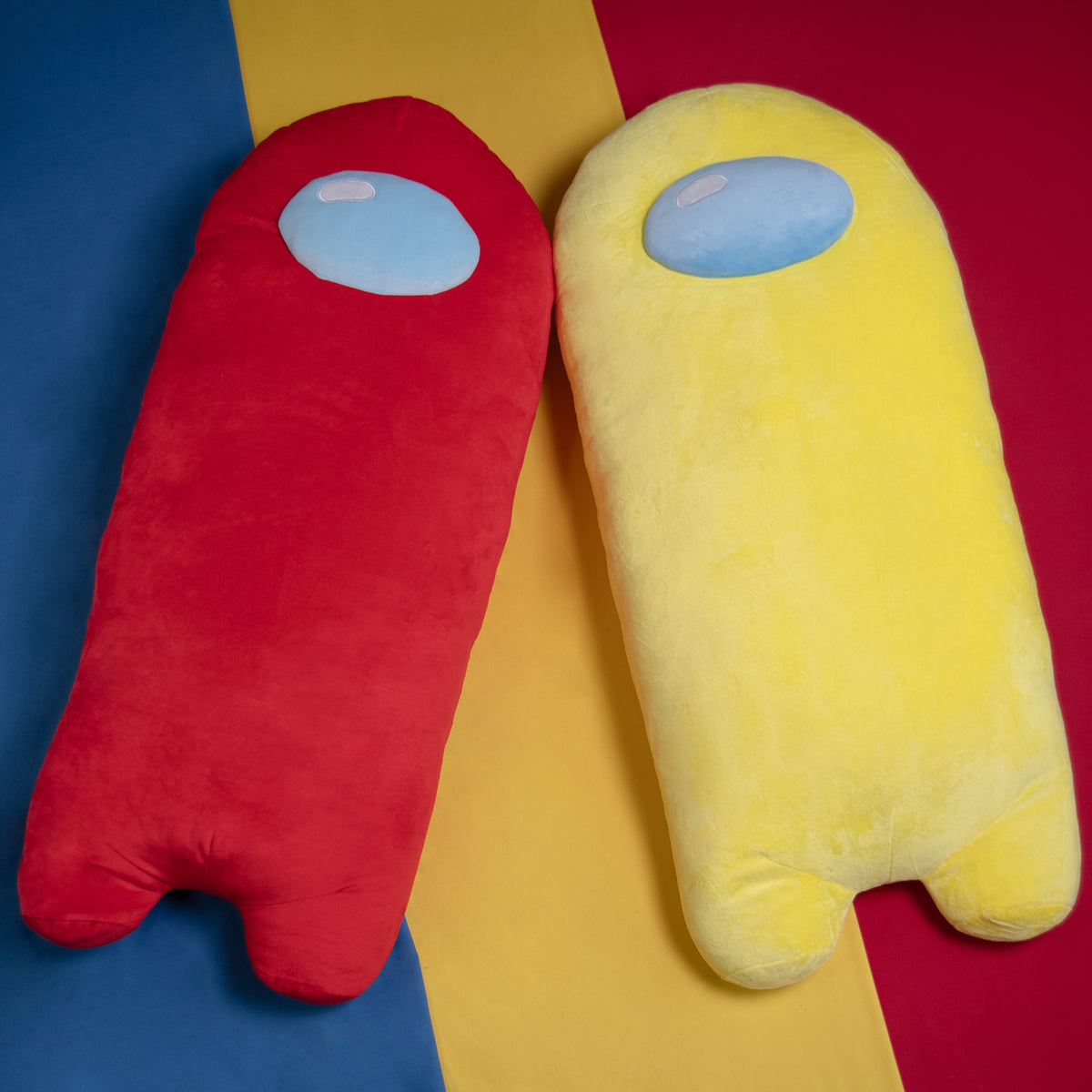 A lifestyle photograph of the red and yellow longbean plushies laying side by side against a blue, yellow, and red background.