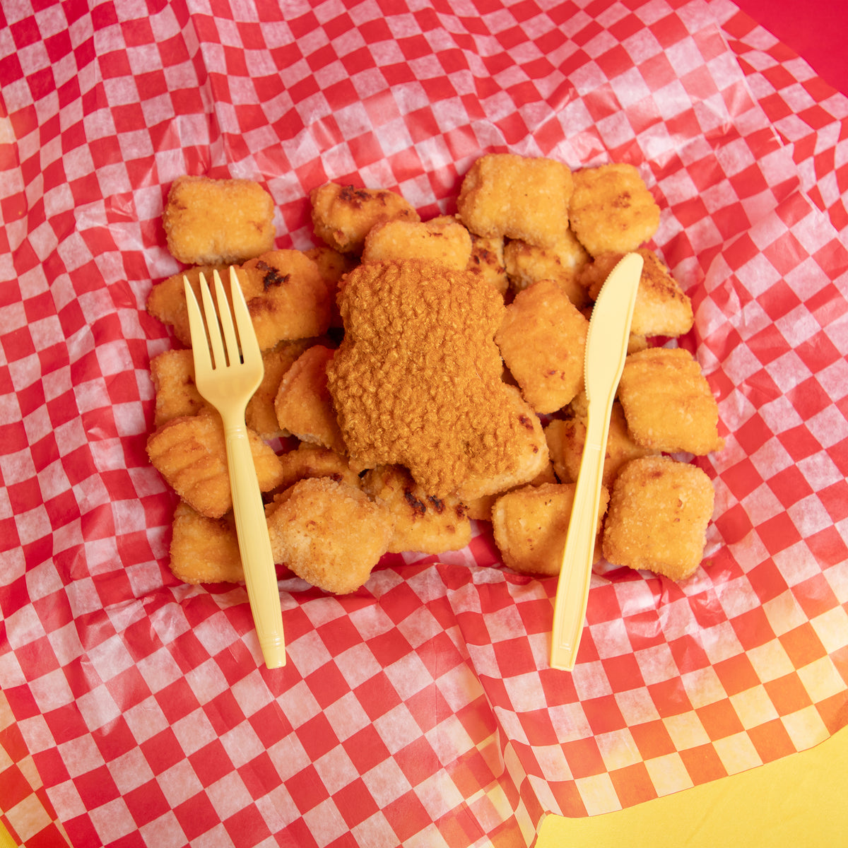 An overhead photograph of the Crewmate chicken nugget plush laying in a pile of real chicken nuggets. There is a plastic fork and knife on either side. All the items are piled on top of checkered red and white diner tissue paper. 