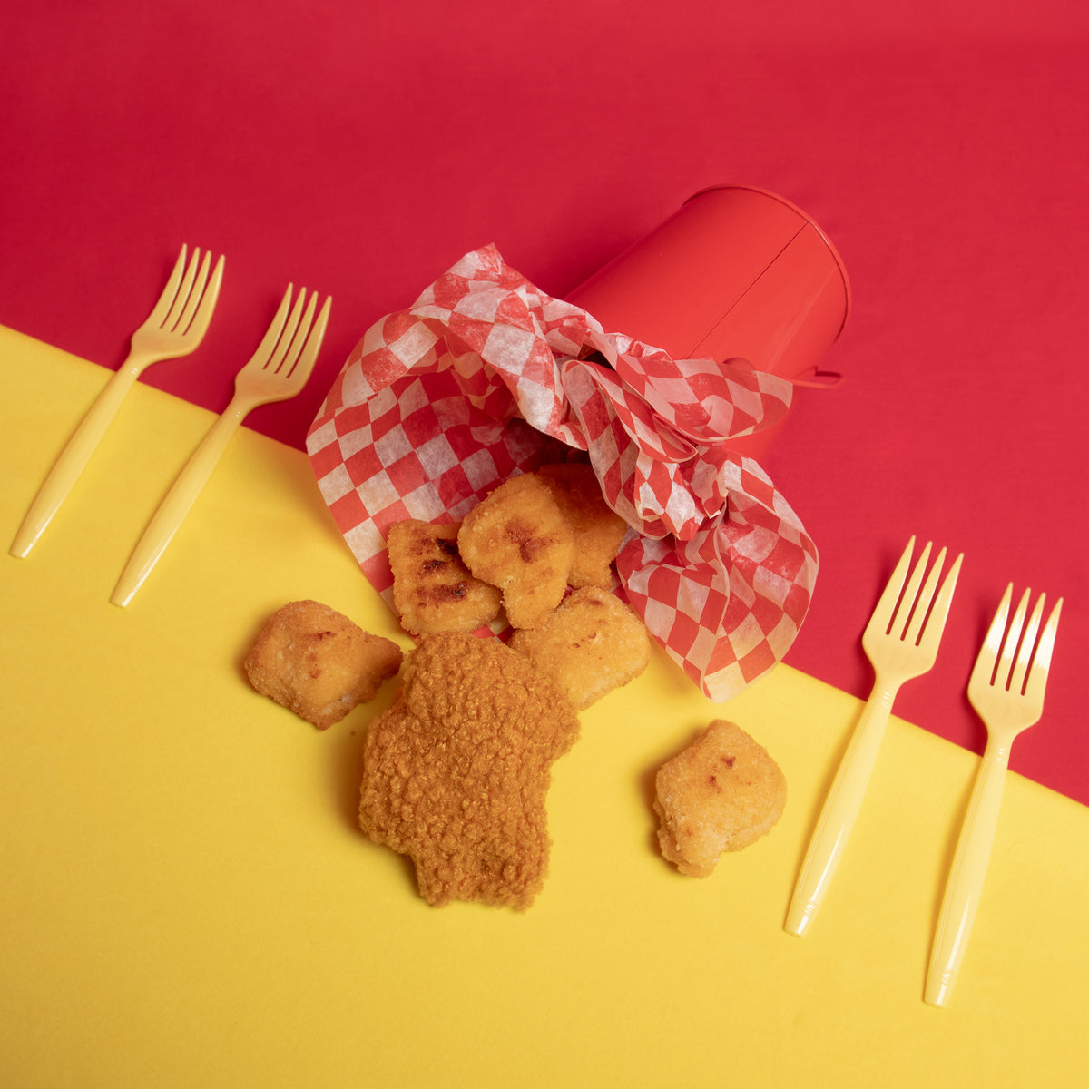 An overhead photograph of a cup lined with checkered tissue paper, real chicken nuggets and the Crewmate chicken nugget spill out of the cup. There are two forks placed on either side of the cup. Photo taken against a red and yellow background. 