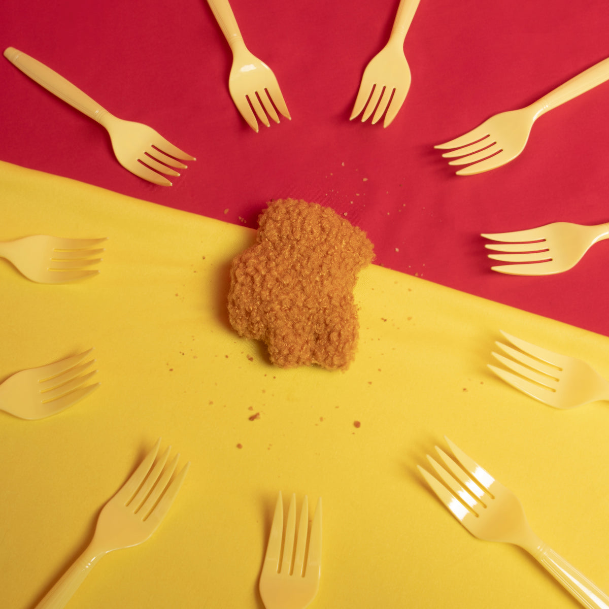 An overhead shot of the Crewmate chicken nugget surrounded by a circle of plastic forks against a red and yellow background. 