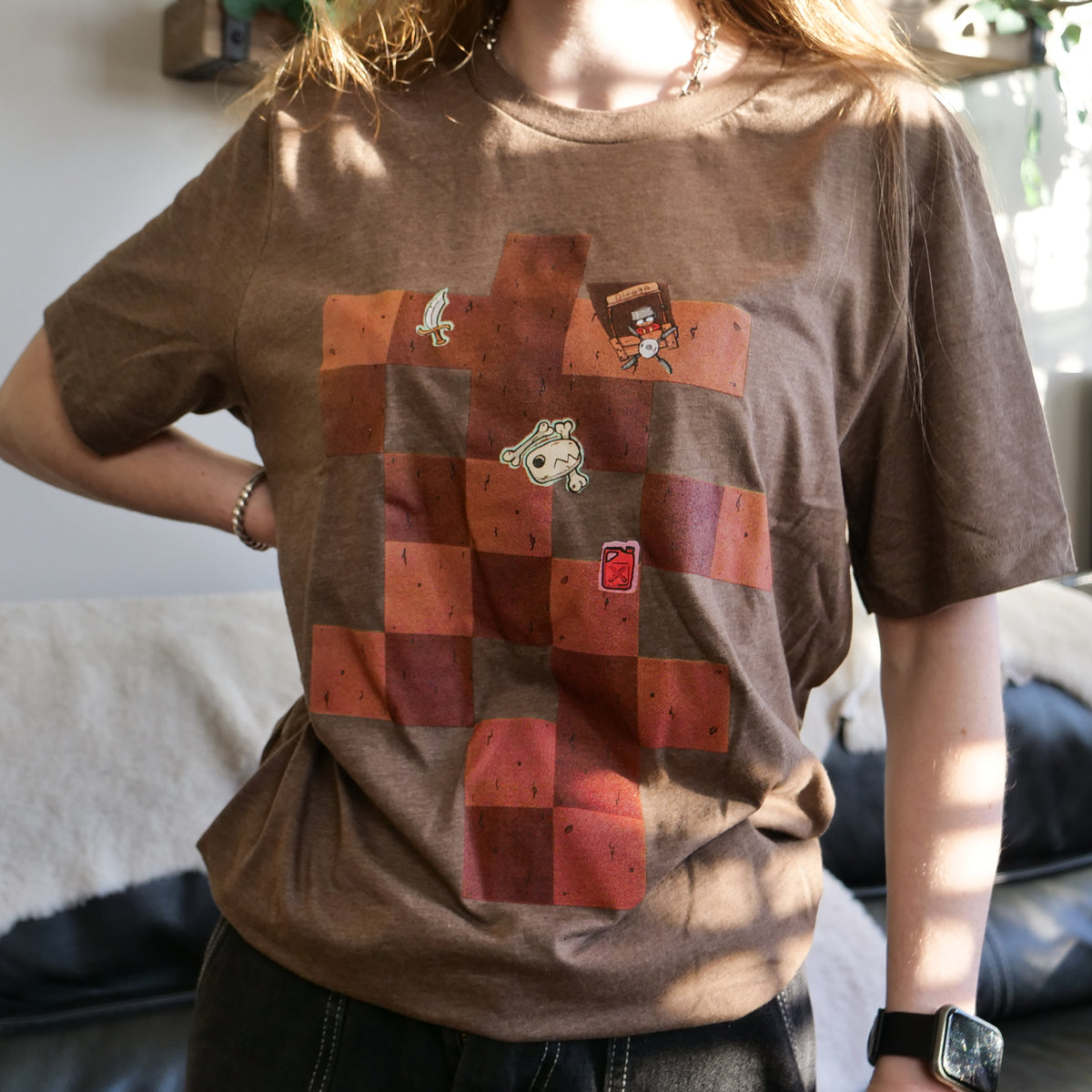 A close-up of someone with long red hair in their living room, wearing the Dig2China Tee, a brown T-shirt with a brown square pattern on it. Scattered on the brown square pattern are a cuirass, an animal skull, a wooden stall saying “Digger” with a person behind it, and a jar of gasoline. The person has their hand at their waist and is wearing an Apple watch, and the image is cropped so that you cannot see the person’s face or anything below their waist. 