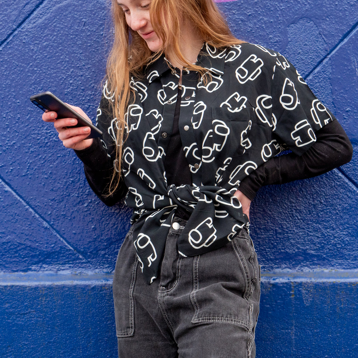 A pale-skinned person with long red hair is holding a phone in front of a blue background with a tiled pattern. They are looking at the phone and smiling. They are wearing grey jeans and a long-sleeved black shirt, and the Beanscape Button-Up (a black short-sleeved button-up covered in crewmates from Among Us outlined in white) tied at the waist on top of the long-sleeved black shirt.