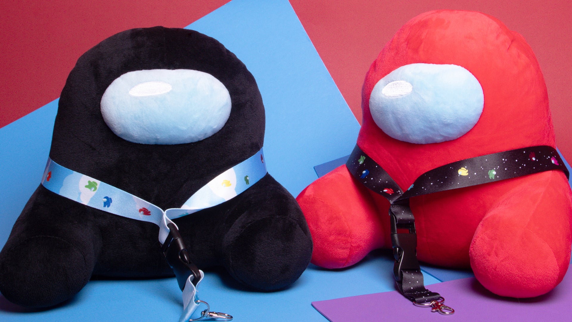 A Black and Red Sitting Crewmate Plush sitting side by side against a red, blue, and purple color blocked background. Each of the Crewmates are wearing a Crewmate lanyard.