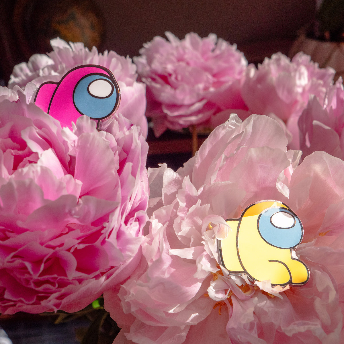 A photo of the yellow and pink Among Us: Mini Crewmate Enamel Pin each laying in the blossoms of pink carnations.