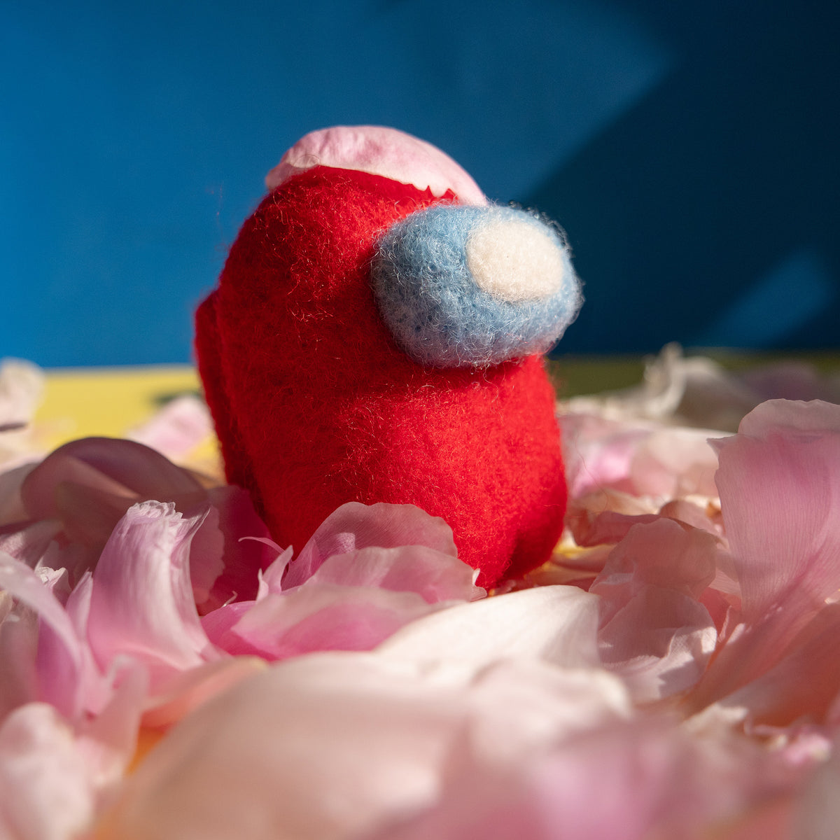 Red felted crewmate standing in a pile of pnk flower petals, with one flower petal on its head