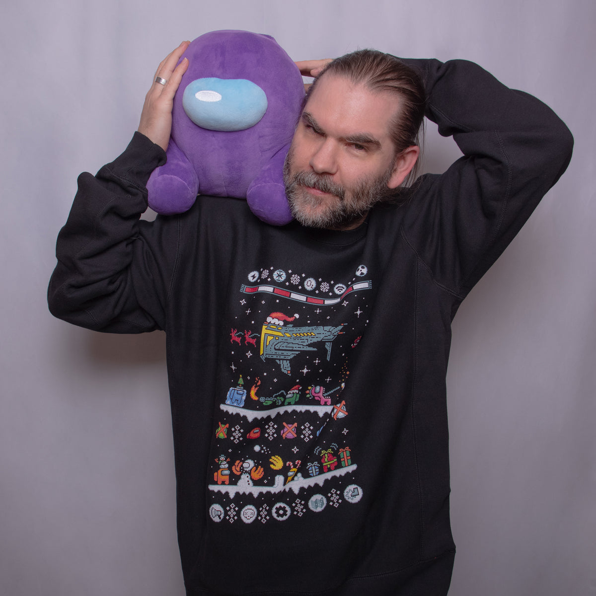 A photograph of a model wearing the  Black Northern Hemisphere longsleeve sweatshirt holding a purple Crewmate plush on their left shoulder. 