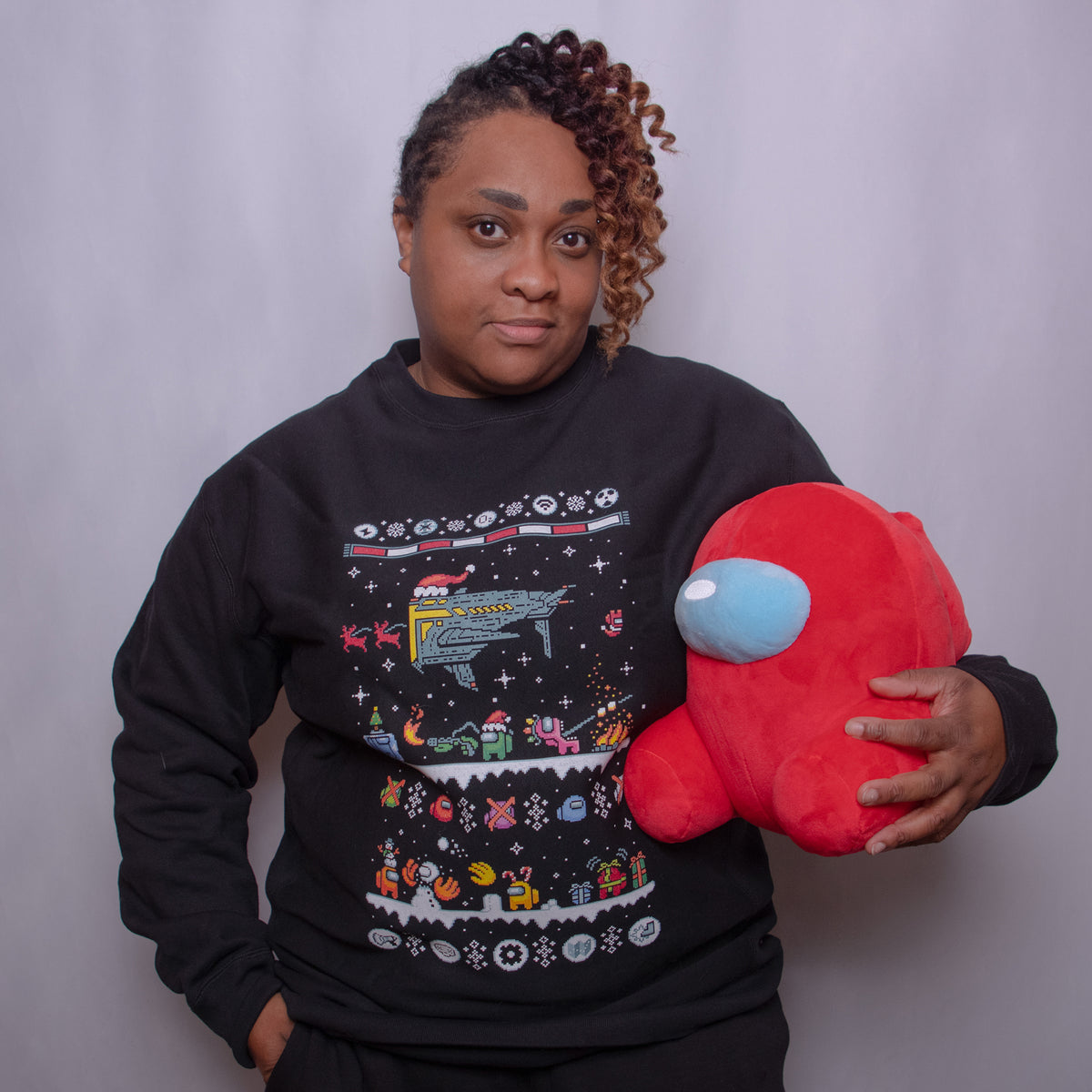A photograph of a model wearing the Black Northern Hemisphere longsleeve sweatshirt holding a red Crewmate plush