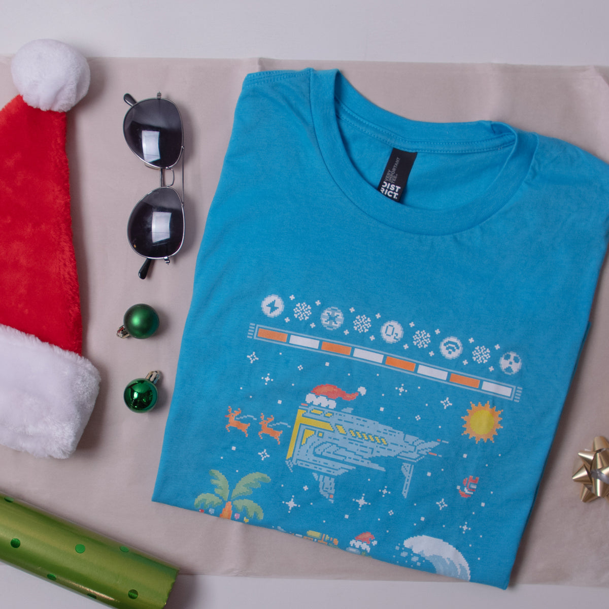 A photograph of the Southern Hemisphere Tee folded and arranged onto a scene next to a santa hat, sunglasses, tiny ornaments, wrapping paper, and a bow. 
