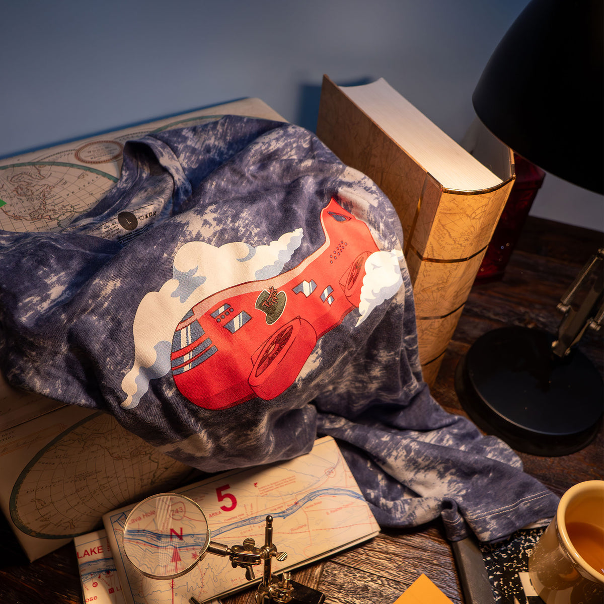 A lifestyle photograph of The Airship Tee. The shirt is sequestered in a corner of a desk draped over a a cartography box. Surrounding the shirt are items relating to adventure such as a folded map, a magnifying glass, a notebook, and a golden hardbound map-covered book. 