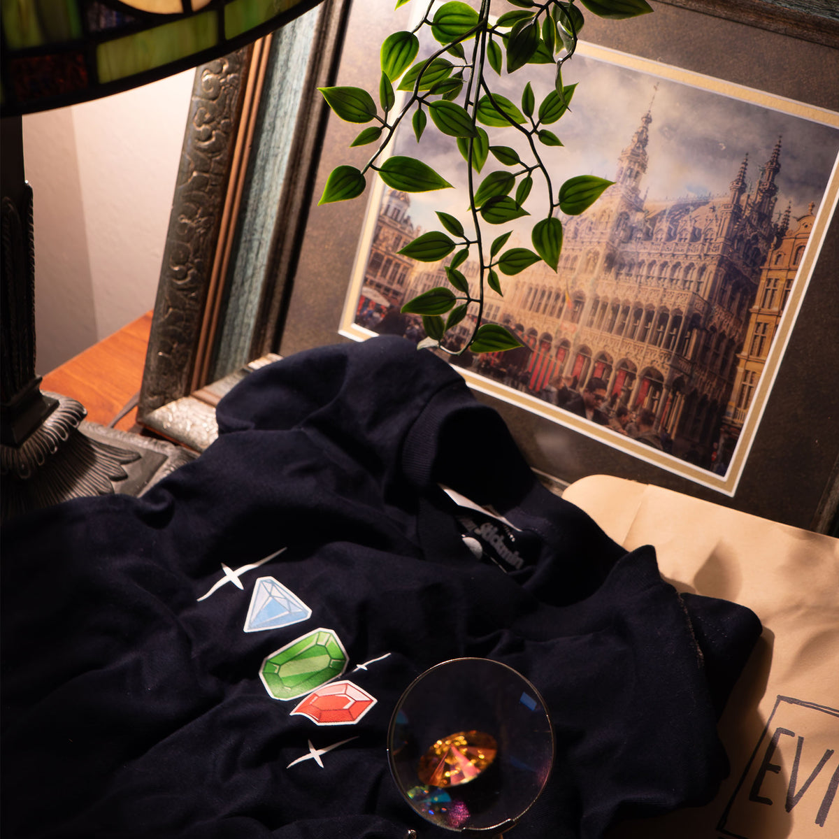 A lifestyle photograph close up of the Jewel Baron Tee. The Tee lies neatly on a desk covering up a manila envelope that says “evidence.” It’s surround by several objects including a framed photograph of an ornate building, a magnifying glass, Tiffany lamp, and plant vines.