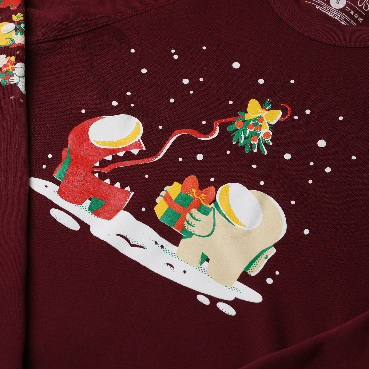 A close up photograph of the partial left sleeve detail and center shirt design. The central illustration depicts a snowy scene of a banana Crewmate holding a wrapped gift while the red Impostor uses its tongue to hold up a mistletoe overhead.