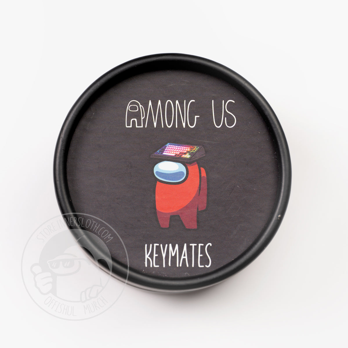 A product photo from above of the black container the keycaps come in. The top of the cardboard cylinder shows a standing red crewmate wearing a rainbow keyboard as a hat. It reads: Among Us: Keymates.”