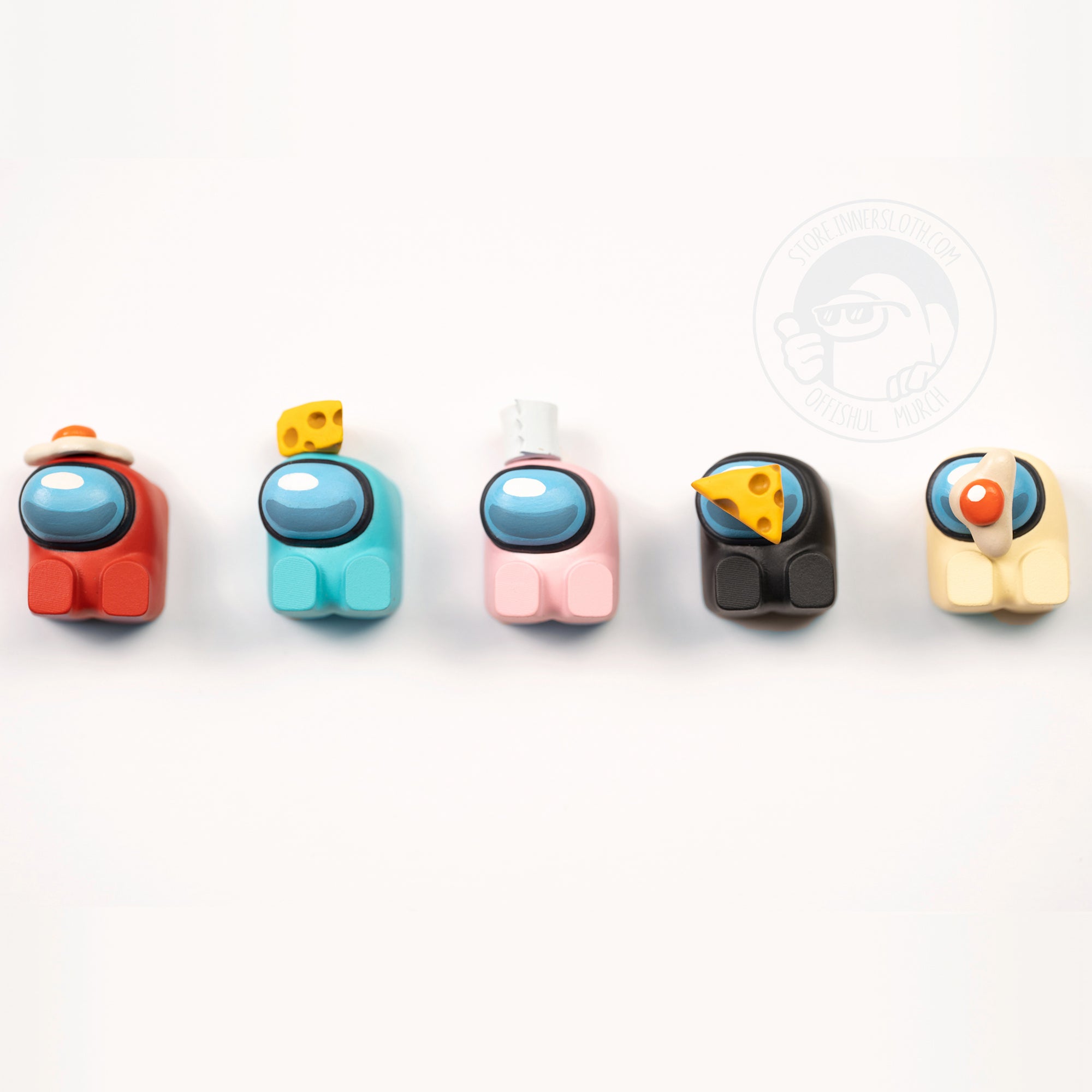 A product photo of five Crewmate keycaps, all in different positions as though they had been dropped together on the table. Colors shown are a black crewmate with no hat laying down, a red crewmate with an egg on its visor laying down, a cyan crewmate with a toilet paper hat sitting up, a pink crewmate sitting up with a cheese hat, and a banana crewmate laying down.