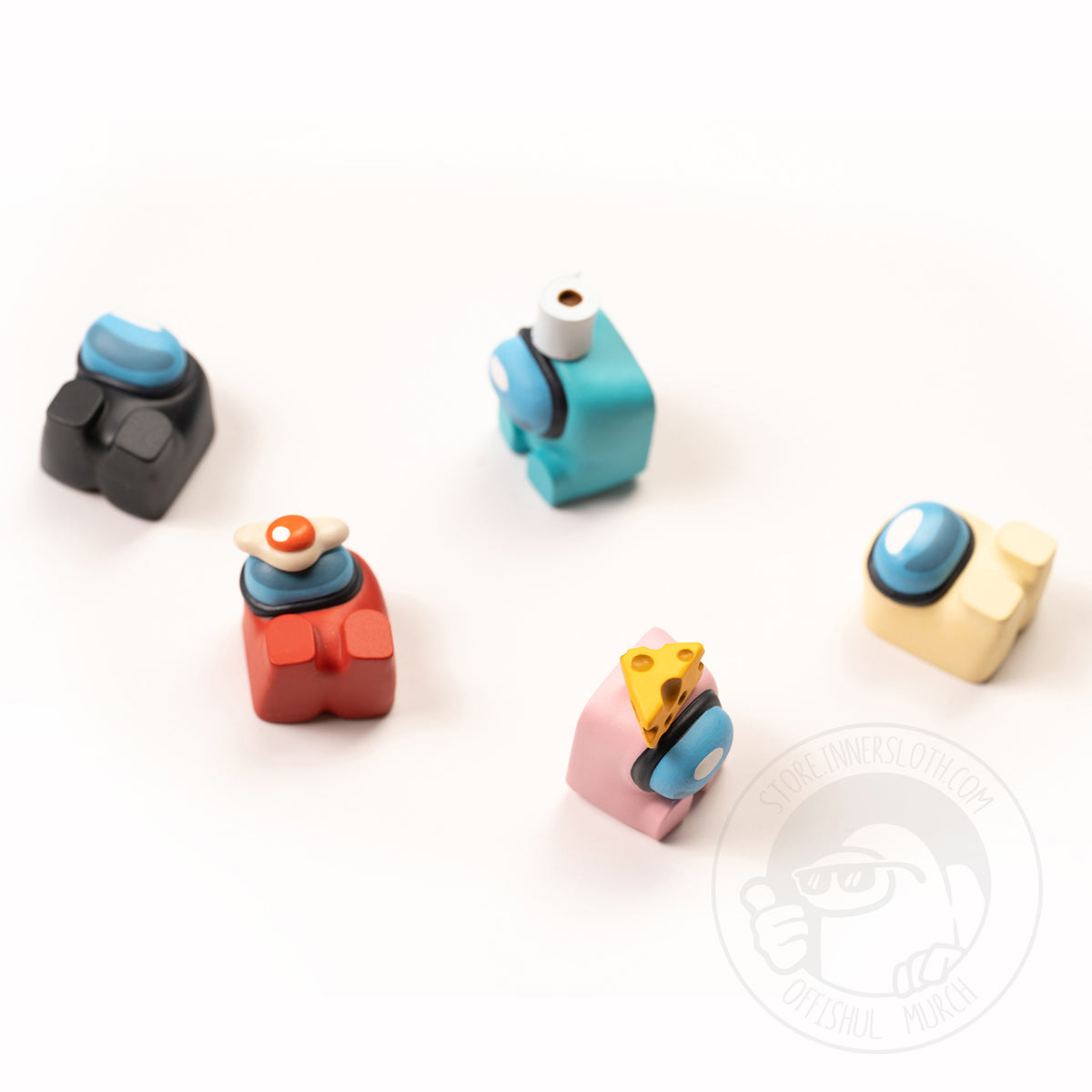 A product photo of five Crewmate keycaps, all in different positions as though they had been dropped together on the table. Colors shown are a black crewmate with no hat laying down, a red crewmate with an egg on its visor laying down, a cyan crewmate with a toilet paper hat sitting up, a pink crewmate sitting up with a cheese hat, and a banana crewmate laying down.