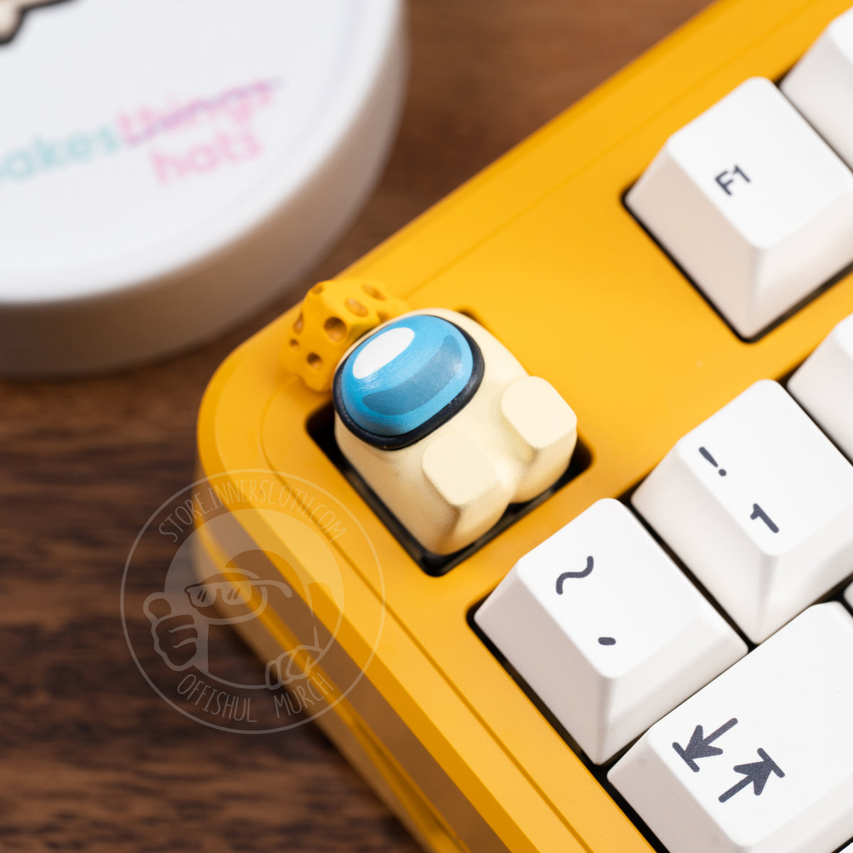 A lifestyle photo of the banana crewmate keycap taking the place of the escape key on a yellow keyboard with white keys. The crewmate keycap figure lays on its back, and is shown with a cheese wedge atop its head.