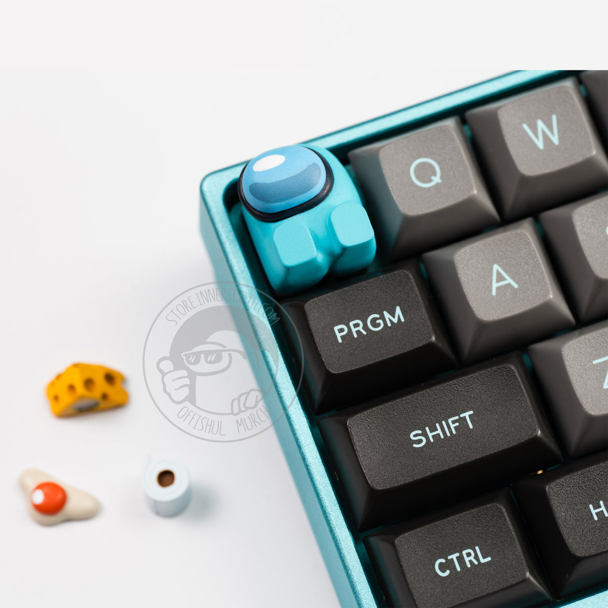 A product photo of the cyan keycap taking the place of the escape key on a cyan keyboard with grey and black keys. The three accessories (fried egg, cheese, and toilet paper) are out of focus on the white table beside the keyboard.